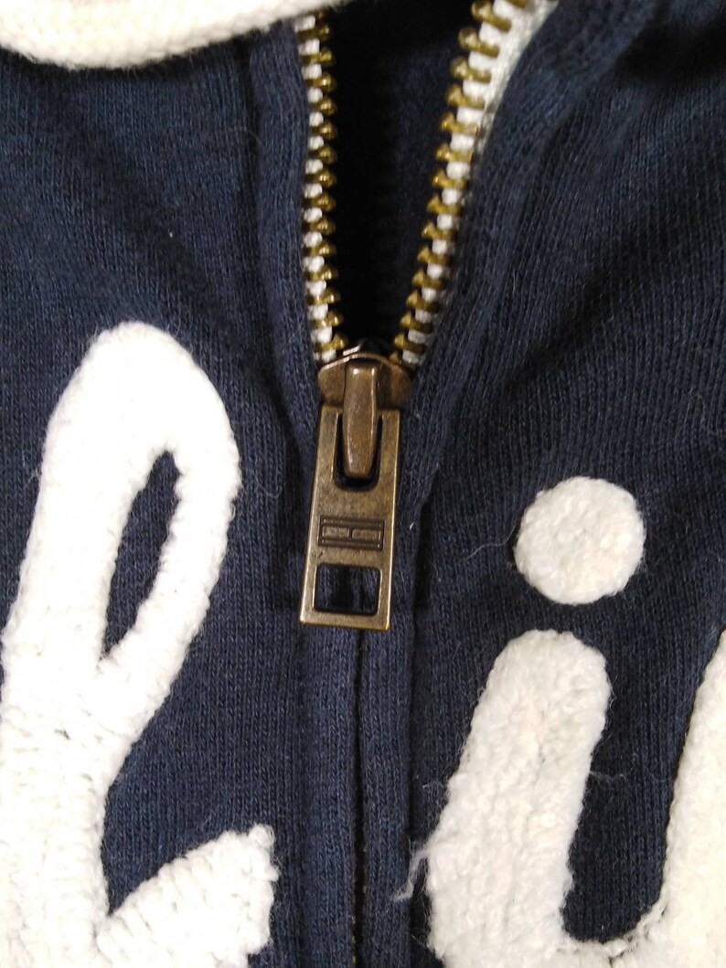 May include: A navy blue zippered hoodie with white embroidered lettering. The zipper is gold metal and has a pull tab.
