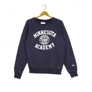May include: A navy blue sweatshirt with a white embroidered logo that says "Minnesota Academy" and a crest with a bird and a star. The sweatshirt has a Champion logo on the left sleeve.
