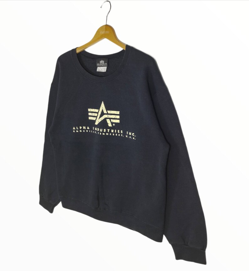 May include: A black Alpha Industries sweatshirt with a gold star logo and the text "ALPHA INDUSTRIES INC. KNOXVILLE, TENNESSEE, U.S.A."