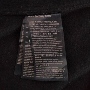 May include: A black clothing label with white text that reads "www.tommy.com" and "MADE IN CHINA/FABRIQU&Eacute; EN CHINE/HECHO EN CHINA/CIN'DE URETILMISTIR/VYROBENO V Č&Iacute;NĚ/WYPRODUKOWANO W CHINACH/DIBUAT DI CINA/صنع في الصين/원산지 : 중국/產地:中國/FABRICADO NA CHINA/" The label also includes information about the fabric composition, which is 60% cotton and 40% polyester.