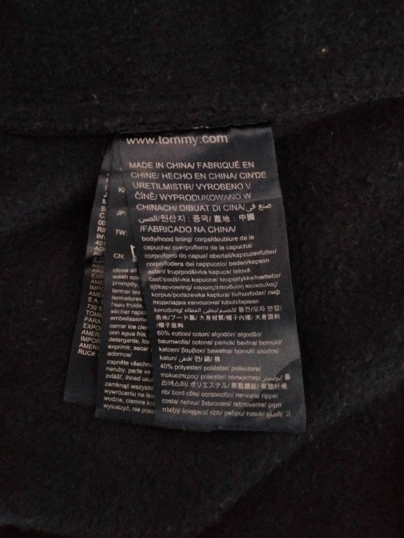 May include: A black clothing label with white text that reads "www.tommy.com" and "MADE IN CHINA/FABRIQU&Eacute; EN CHINE/HECHO EN CHINA/CIN'DE URETILMISTIR/VYROBENO V Č&Iacute;NĚ/WYPRODUKOWANO W CHINACH/DIBUAT DI CINA/صنع في الصين/원산지 : 중국/產地:中國/FABRICADO NA CHINA/" The label also includes information about the fabric composition, which is 60% cotton and 40% polyester.