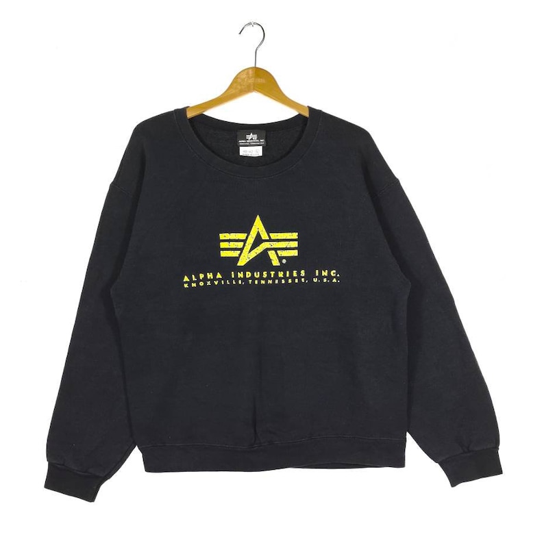 May include: Black sweatshirt with a yellow Alpha Industries logo and the text "ALPHA INDUSTRIES INC. KNOXVILLE, TENNESSEE, U.S.A."