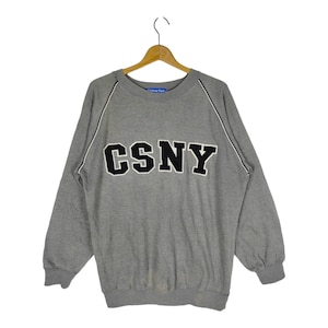 Könnte beinhalten: Grauer Sweatshirt mit einem schwarzen und weißen "CSNY"-Logo auf der Vorderseite. Der Sweatshirt hat weiße Paspeln an den Schultern und Ärmeln.