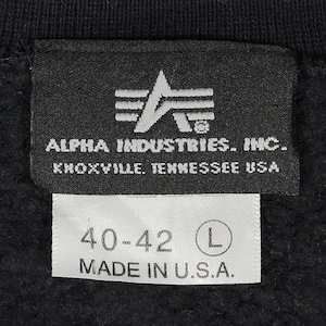 May include: Black and white label with the text "ALPHA INDUSTRIES. INC. KNOXVILLE. TENNESSEE USA" and a logo of a stylized letter A with three stripes. A white label below reads "40-42 L MADE IN U.S.A."