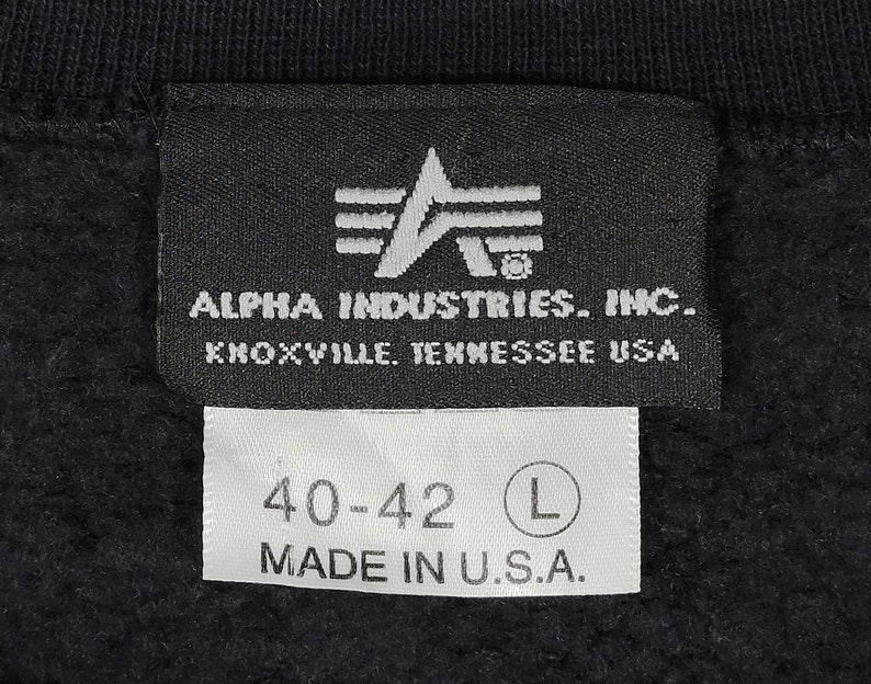 May include: Black and white label with the text "ALPHA INDUSTRIES. INC. KNOXVILLE. TENNESSEE USA" and a logo of a stylized letter A with three stripes. A white label below reads "40-42 L MADE IN U.S.A."