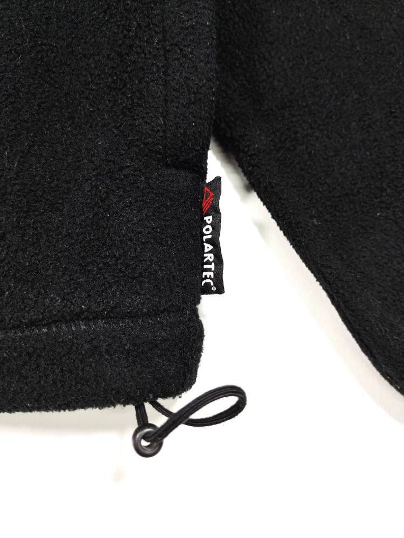 K&ouml;nnte beinhalten: Schwarze Fleecejacke mit schwarzem Kordelzug und rotem "POLARTEC"-Etikett.