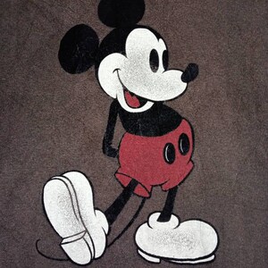 May include: A brown t-shirt featuring a black and white image of Mickey Mouse. Mickey Mouse is wearing red shorts and white shoes. The text "Mickey Mouse" is printed below the image.