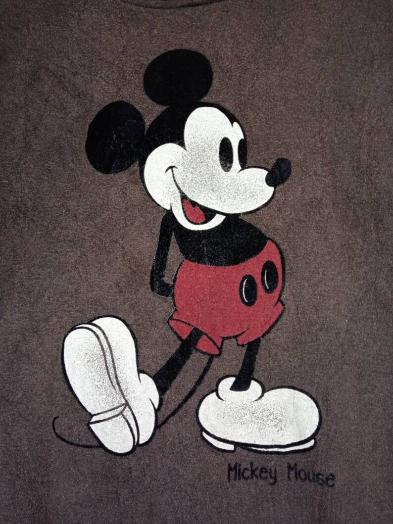May include: A brown t-shirt featuring a black and white image of Mickey Mouse. Mickey Mouse is wearing red shorts and white shoes. The text "Mickey Mouse" is printed below the image.