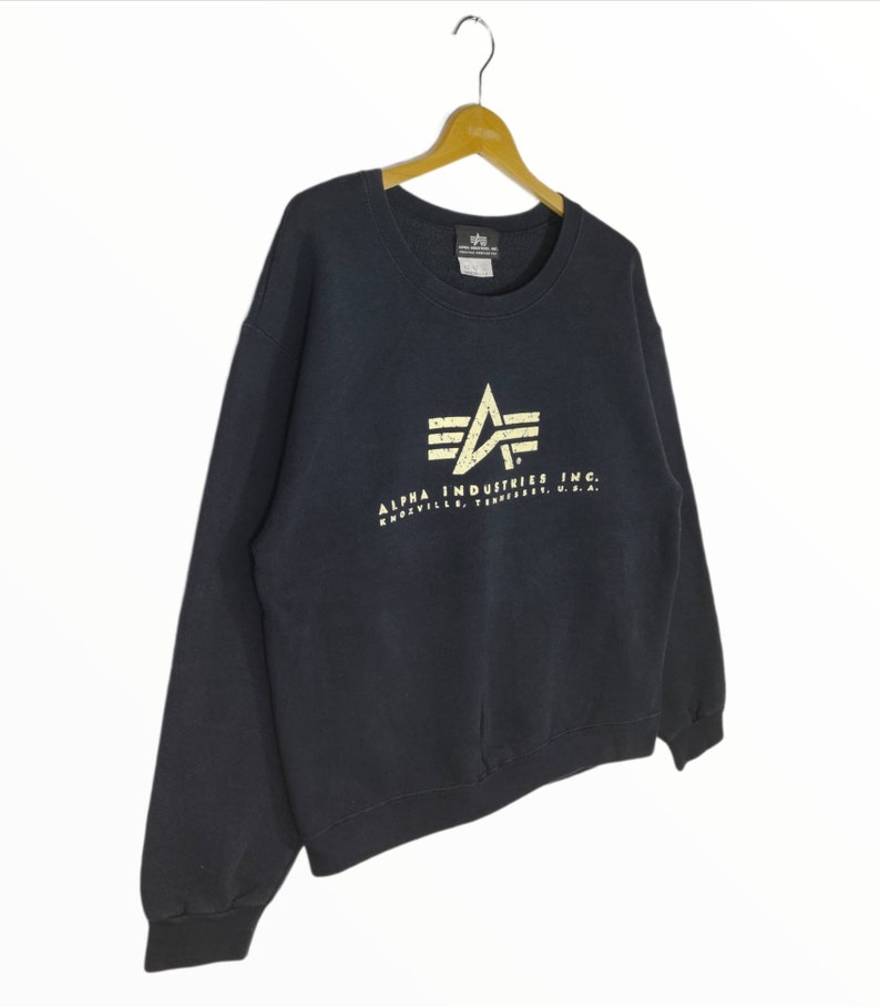 May include: A dark blue Alpha Industries sweatshirt with a white logo featuring a stylized airplane wing and the text "Alpha Industries Inc. Knoxville, Tennessee, U.S.A."