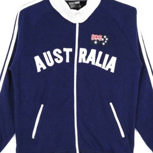 May include: A navy blue zippered jacket with white stripes down the sleeves and a white embroidered "AUSTRALIA" across the chest. The jacket has a small embroidered Australian flag on the right side of the chest.