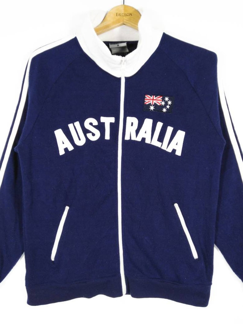 May include: A navy blue zippered jacket with white stripes down the sleeves and a white embroidered "AUSTRALIA" across the chest. The jacket has a small embroidered Australian flag on the right side of the chest.