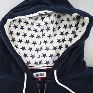May include: A navy blue zip-up hoodie with a white star pattern lining the hood. The hoodie has a white embroidered logo that says "Hilfiger Denim" and a white drawstring. The tag says "An American Original".