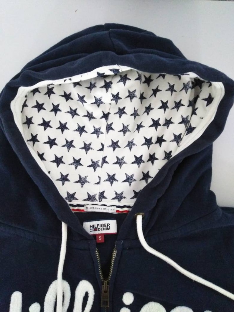 May include: A navy blue zip-up hoodie with a white star pattern lining the hood. The hoodie has a white embroidered logo that says "Hilfiger Denim" and a white drawstring. The tag says "An American Original".