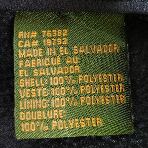 K&ouml;nnte beinhalten: Ein gr&uuml;nes Stofflabel mit gelbem Text. Das Etikett enth&auml;lt den Text "RN# 76382", "CA# 19792", "MADE IN EL SALVADOR", "FABRIQU&Eacute; AU EL SALVADOR", "SHELL: 100% POLYESTER", "VESTE 100% POLYESTER", "LINING 100% POLYESTER", "DOUBLURE: 100% POLYESTER".