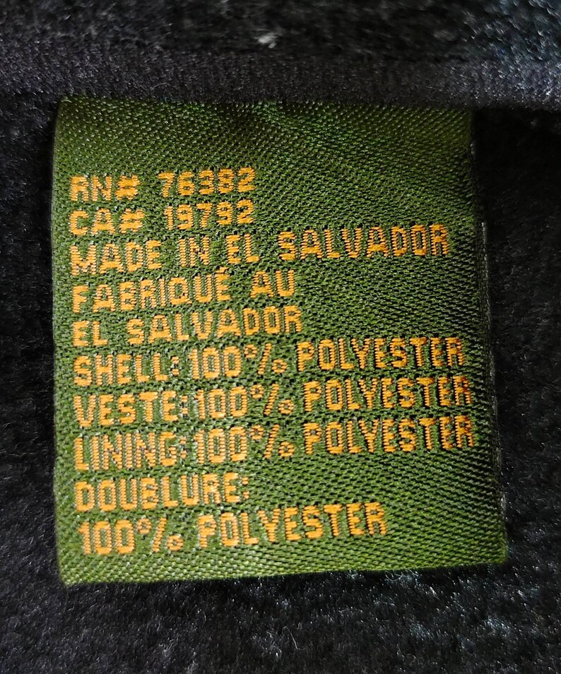 K&ouml;nnte beinhalten: Ein gr&uuml;nes Stofflabel mit gelbem Text. Das Etikett enth&auml;lt den Text "RN# 76382", "CA# 19792", "MADE IN EL SALVADOR", "FABRIQU&Eacute; AU EL SALVADOR", "SHELL: 100% POLYESTER", "VESTE 100% POLYESTER", "LINING 100% POLYESTER", "DOUBLURE: 100% POLYESTER".
