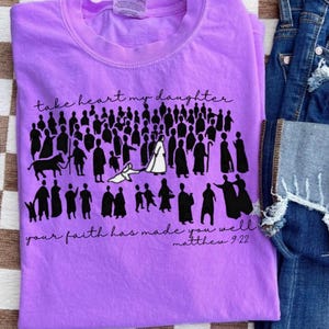 May include: A lilac-colored t-shirt with the text "take heart my daughter" and "your faith has made you well matthew 9:22" in black script. The shirt features a graphic of silhouetted figures.