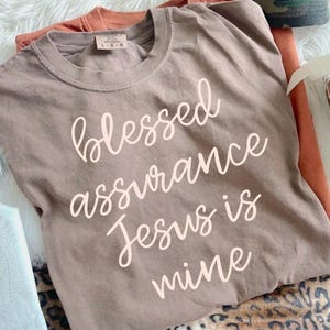 May include: A light brown t-shirt with the words "blessed assurance Jesus is mine" in a cursive font. The shirt is folded with an orange shirt underneath. A camouflage baseball cap is visible in the background.