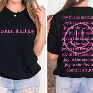 Puede incluir: Una camiseta negra con texto rosa y un gráfico de cara sonriente. El frente muestra una cara sonriente y las palabras count it all joy. La parte posterior presenta una gran cara sonriente rodeada por el texto joy in the morning, evening, waiting, knowing, laughing, weeping, breaking, mending, seeking, finding y count it all JOY.
