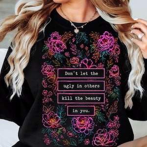 May include: A black long-sleeve sweatshirt featuring a vibrant floral print in shades of neon pink, purple, and teal. The center displays the text: Don't let the ugly in others kill the beauty in you, framed by colorful botanical illustrations.