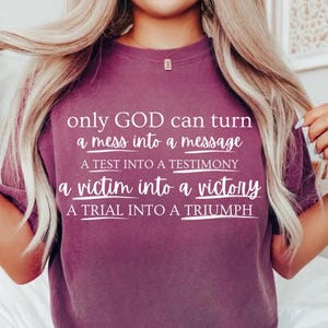 May include: A plum-coloured short-sleeve t-shirt featuring white text that reads: only GOD can turn a mess into a message, A TEST INTO A TESTIMONY, a victim into a victory, A TRIAL INTO A TRIUMPH, with each line underlined by a white graphic stroke.