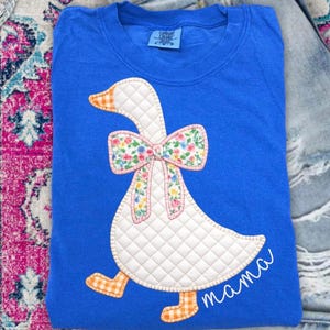 May include: A bright blue t-shirt featuring a quilted white goose appliqué with an orange and white checkered beak and feet. The goose wears a floral print bow and the word mama is embroidered in white cursive script on the lower right side.