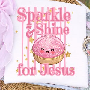 May include: White t-shirt with a pink design. The design includes the words "Sparkle & Shine for Jesus" in pink, vertical pink stripes, and a cute pink dessert with a smiling face and gold glitter accents.