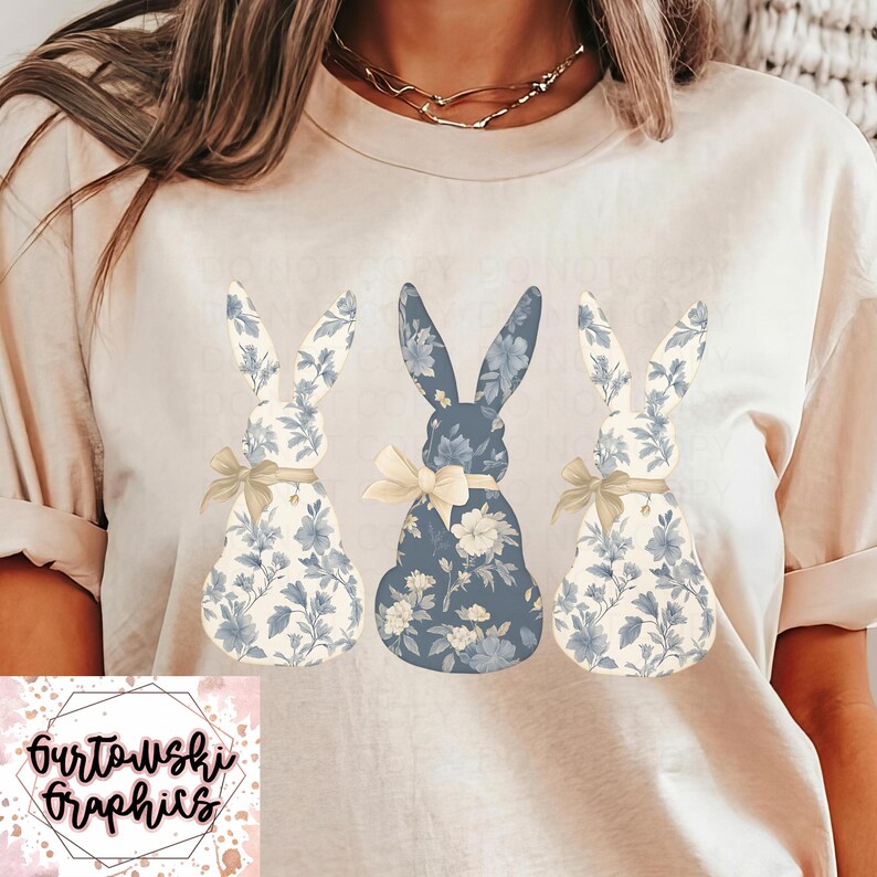 May include: A white t-shirt with three blue and white floral bunny rabbits printed on the front. The bunnies are arranged in a row, with the middle bunny slightly larger than the other two. The bunnies have a light brown bow tied around their necks.