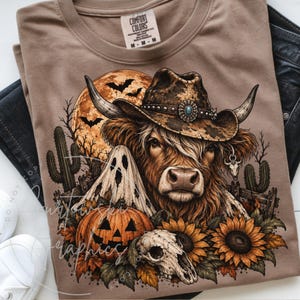 May include: A tan t-shirt featuring a graphic of a Highland cow wearing a cowboy hat, a ghost, a jack-o'-lantern, sunflowers, a skull, and cacti against a full moon background. The inner neck label reads Comfort Colors, 100% cotton, made in Honduras.