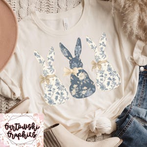 May include: A white t-shirt with three blue and white floral bunny silhouettes. The bunnies are arranged in a row, with the middle bunny slightly larger than the others. The bunnies have a light yellow bow tied around their necks.