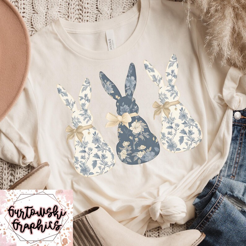 May include: A white t-shirt with three blue and white floral bunny silhouettes. The bunnies are arranged in a row, with the middle bunny slightly larger than the others. The bunnies have a light yellow bow tied around their necks.
