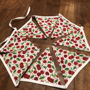May include: A white fabric bunting with red cherries and green leaves on a white background. The bunting is made of triangles with white trim and is strung together with white ribbon.