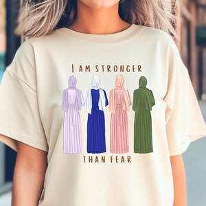 Peut inclure: Un t-shirt beige clair avec un graphique de cinq femmes portant des foulards et des robes longues. Le texte "Je suis plus forte que la peur" est imprim&eacute; sous le graphique.