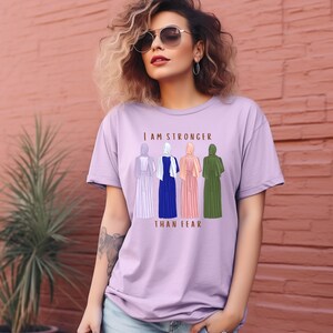 Peut inclure: Un t-shirt lavande avec le texte "Je suis plus forte que la peur" imprim&eacute; sur le devant. Le texte est en lettres brunes et est entour&eacute; d'une illustration de cinq femmes portant des foulards.