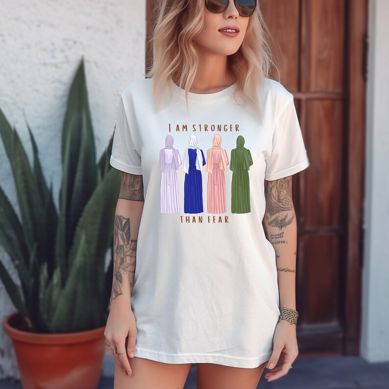 Peut inclure: T-shirt blanc avec un motif de quatre femmes portant des foulards et des robes. Le texte "Je suis plus forte que la peur" est imprim&eacute; sous le motif.