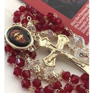 Puede incluir: Un rosario dorado con cuentas de cristal rojo y un crucifijo dorado con una representación de Jesucristo. El rosario también incluye un pequeño medallón dorado con una representación de Jesucristo.