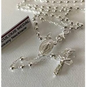 Puede incluir: Un collar de rosario plateado con un crucifijo y un colgante ovalado con una figura religiosa. El collar está hecho de pequeñas cuentas redondas e incluye un cierre. Una etiqueta con la inscripción "Miraculous Medal Rosary Necklace" es visible.