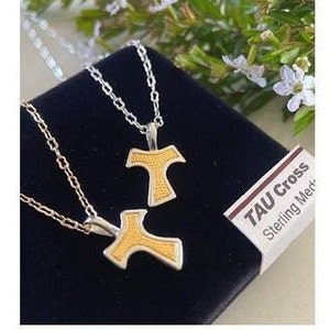Puede incluir: Dos collares de plata con cruces Tau doradas. Los collares se muestran sobre un fondo de terciopelo negro. El texto "TAU Cross Sterling Merk" es visible en una etiqueta blanca.