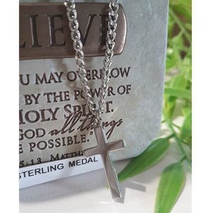 Puede incluir: Un collar de cadena de plata con un colgante de cruz de plata. El colgante tiene un acabado liso y pulido. El collar est&aacute; sobre un fondo blanco con una etiqueta que dice "Sterling Medal" y una cita sobre la fe.
