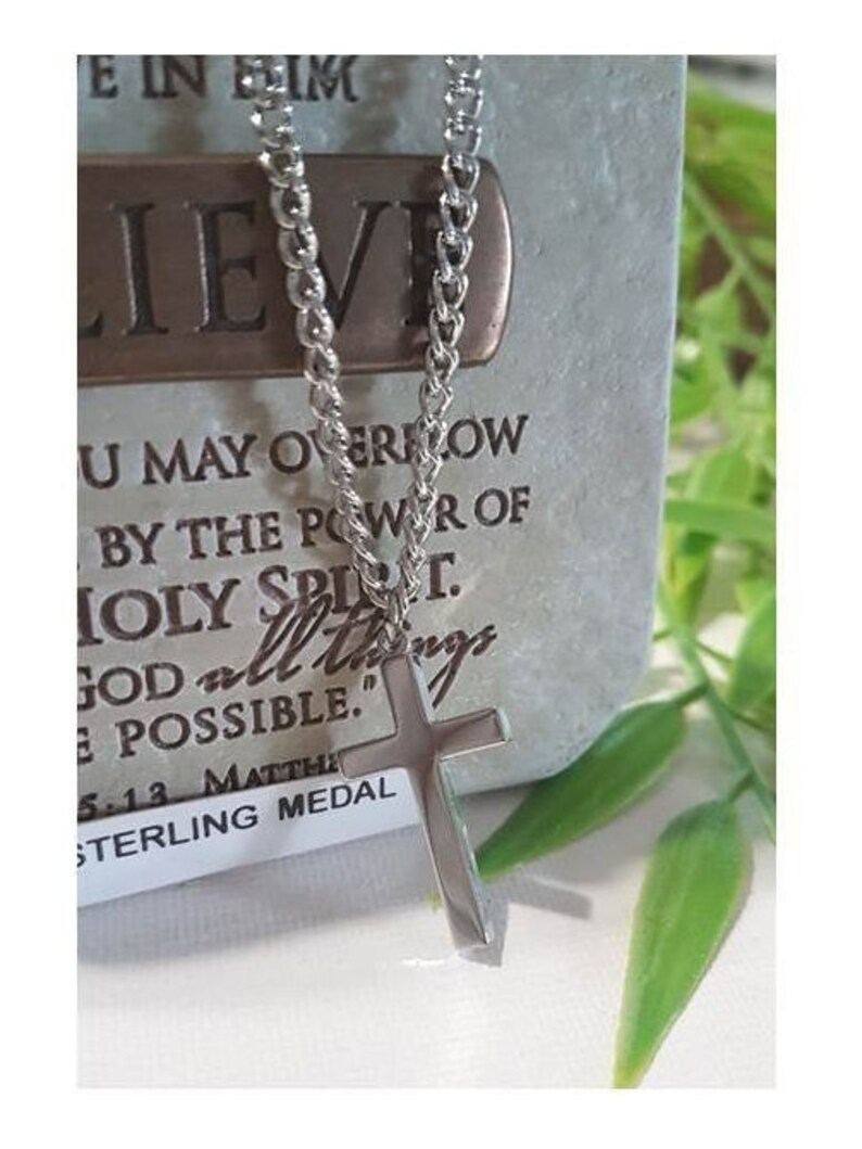 Puede incluir: Un collar de cadena de plata con un colgante de cruz de plata. El colgante tiene un acabado liso y pulido. El collar est&aacute; sobre un fondo blanco con una etiqueta que dice "Sterling Medal" y una cita sobre la fe.