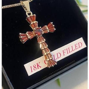 Puede incluir: Un collar con un colgante de cruz de oro relleno, adornado con acentos de piedras preciosas rojas. El colgante está en una caja de terciopelo negro con una etiqueta que dice "18K Gold Filled".