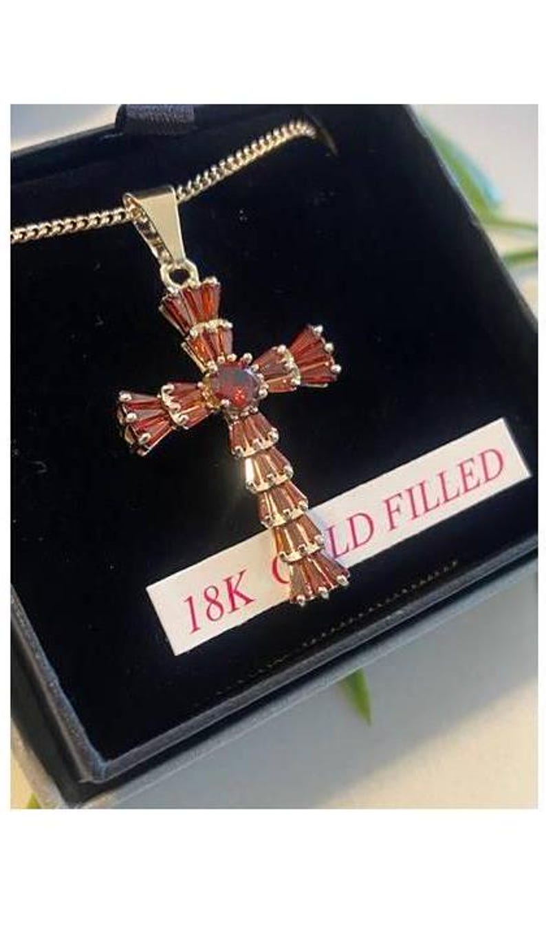Op de afbeelding: Een gold-filled kruis hanger ketting met rode edelsteen accenten. De hanger zit in een zwarte fluwelen doos met een label met de tekst "18K Gold Filled".
