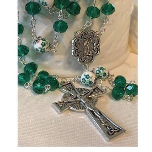 May include: A silver rosary with green and white beads. The centerpiece is a silver Celtic cross with a figure of Jesus Christ. The rosary also features a silver medal with a religious image.