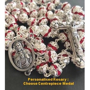 Puede incluir: Un rosario de plata con cuentas de cristal rojo y transparente. El rosario presenta un crucifijo de plata y una medalla de plata con una imagen de Jesús. El texto "Rosario personalizado: Elige la medalla central" es visible en la parte inferior de la imagen.