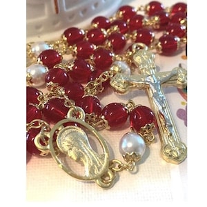 May include: A red and gold rosary with a silver crucifix and a gold charm of the Virgin Mary. The rosary beads are made of glass and the crucifix and charm are made of metal.