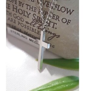 Puede incluir: Un collar de cadena de plata con un colgante de cruz de plata. El collar est&aacute; sobre una superficie marr&oacute;n con el texto "AT YOU MAY OVERFLOW HOPE BY THE POWER OF THE HOLY SPIRIT. ALL THINGS ARE POSSIBLE.  15:18. MATTHEW STERLING MELO 26" impreso en &eacute;l.