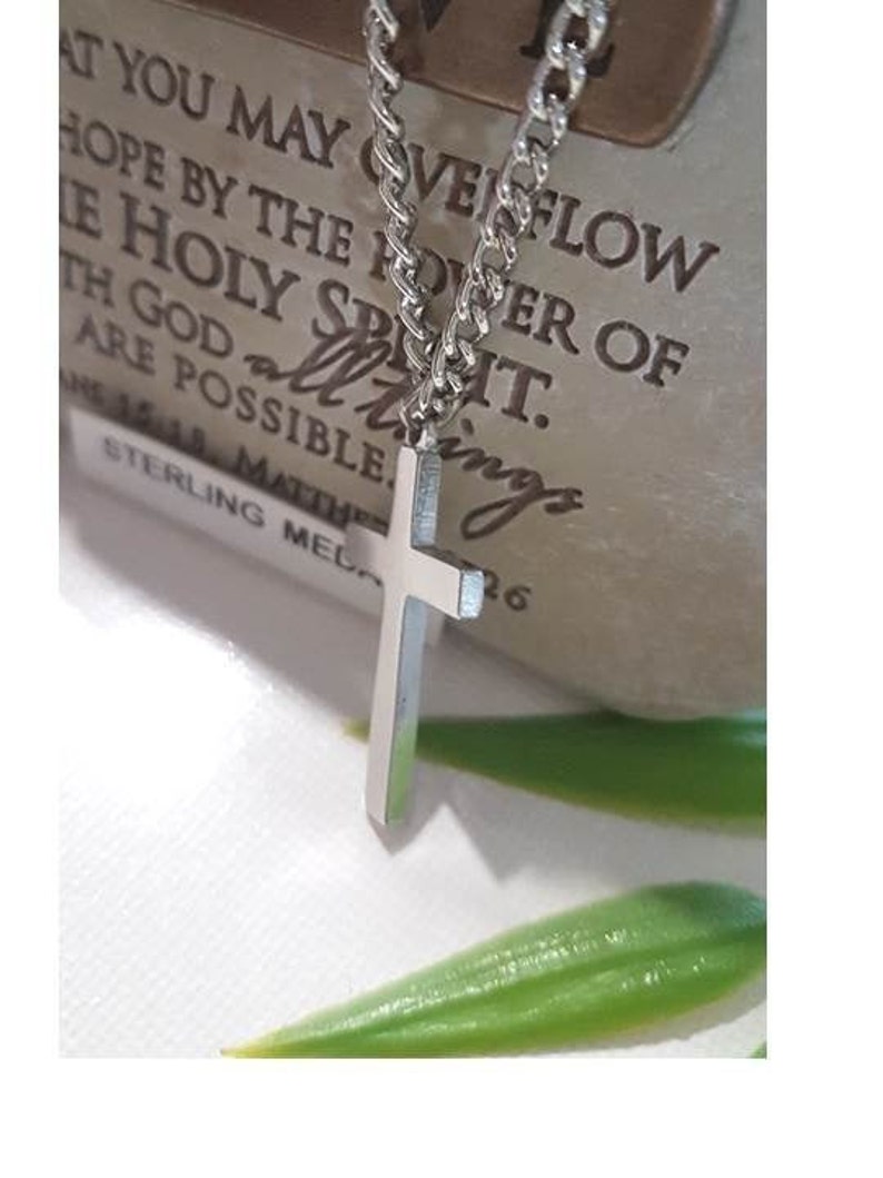 Puede incluir: Un collar de cadena de plata con un colgante de cruz de plata. El collar est&aacute; sobre una superficie marr&oacute;n con el texto "AT YOU MAY OVERFLOW HOPE BY THE POWER OF THE HOLY SPIRIT. ALL THINGS ARE POSSIBLE.  15:18. MATTHEW STERLING MELO 26" impreso en &eacute;l.