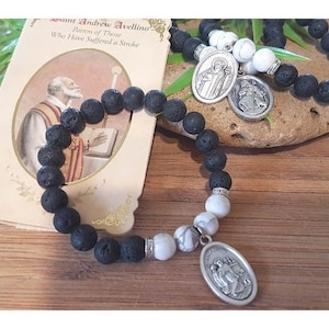 May include: Black and white beaded bracelet with two silver charms featuring religious figures. The bracelet is on a wooden surface with a card that reads "Saint Andrew Avellino Patron of Those Who Have Suffered a Stroke".