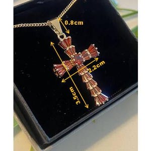 Op de afbeelding: Een gouden kruis hanger met rode edelsteen accenten. De hanger is 3,5 cm hoog en 2,2 cm breed. De hanger is bevestigd aan een gouden ketting.