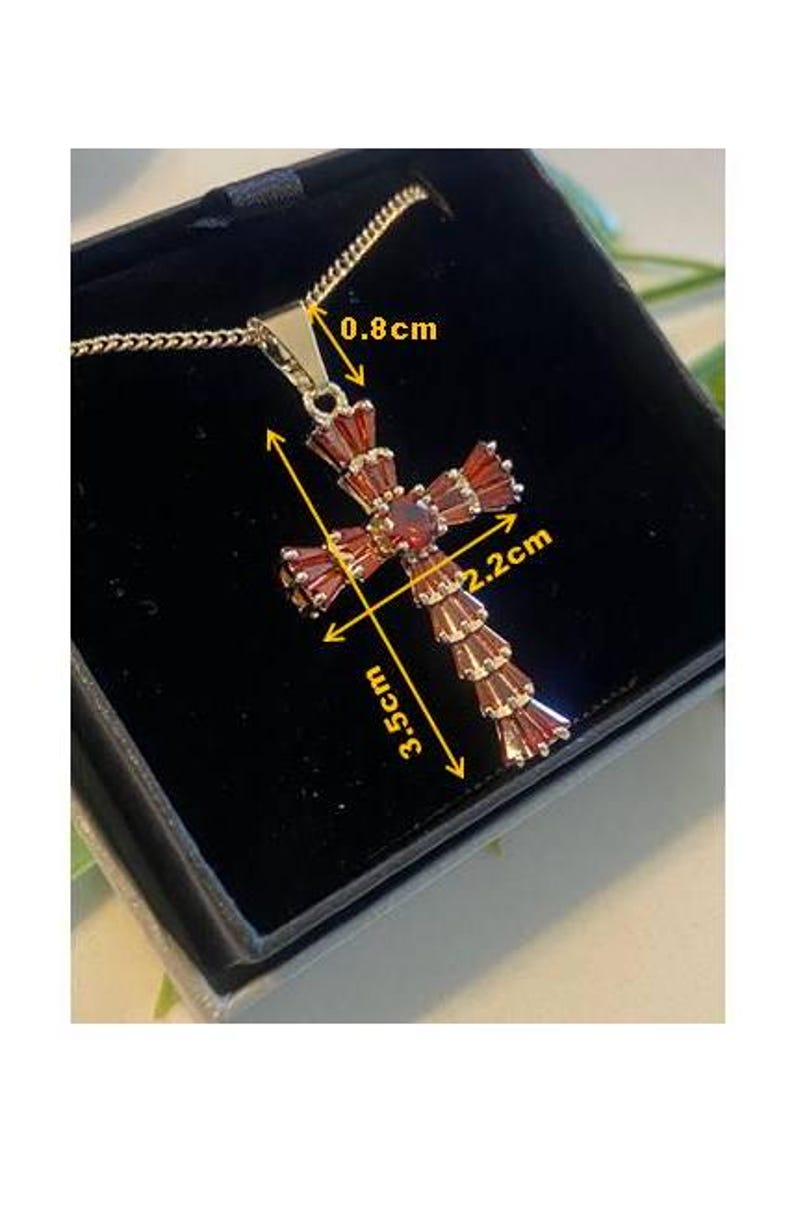 Op de afbeelding: Een gouden kruis hanger met rode edelsteen accenten. De hanger is 3,5 cm hoog en 2,2 cm breed. De hanger is bevestigd aan een gouden ketting.