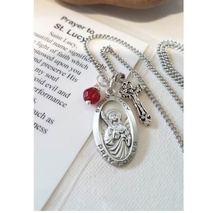 May include: Silver necklace with a silver oval pendant featuring a depiction of Saint Lucy. The pendant has the words "Saint Lucy" and "Pray for Us" inscribed on it. The necklace also includes a small silver cross charm and a red glass bead.
